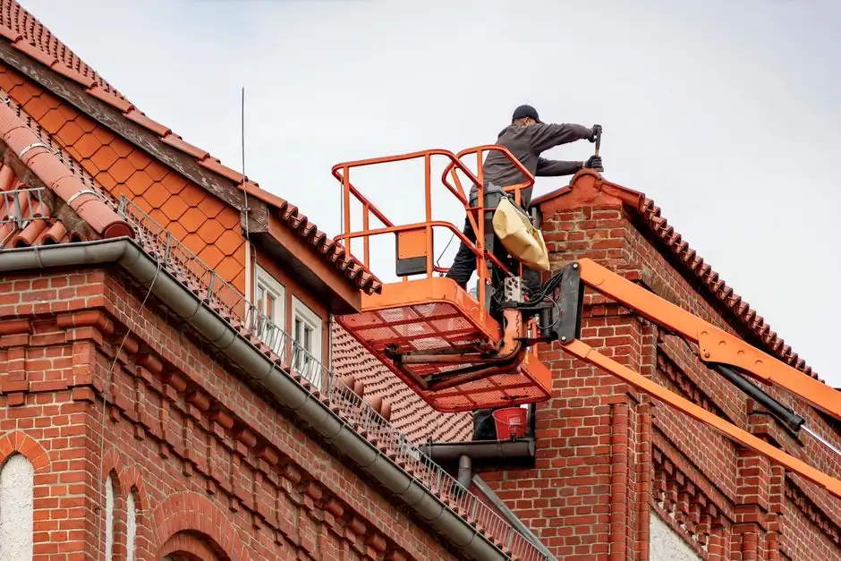 Roofer in a cherry picker conducting maintenance on a red brick building with a tiled roof.